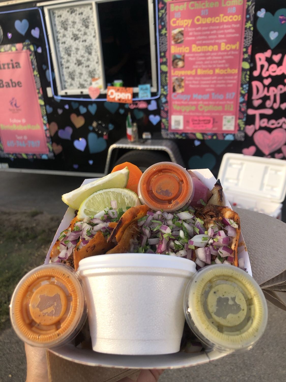 A platter of birria tacos with consome cup and salsas in front of the Birria Babe truck